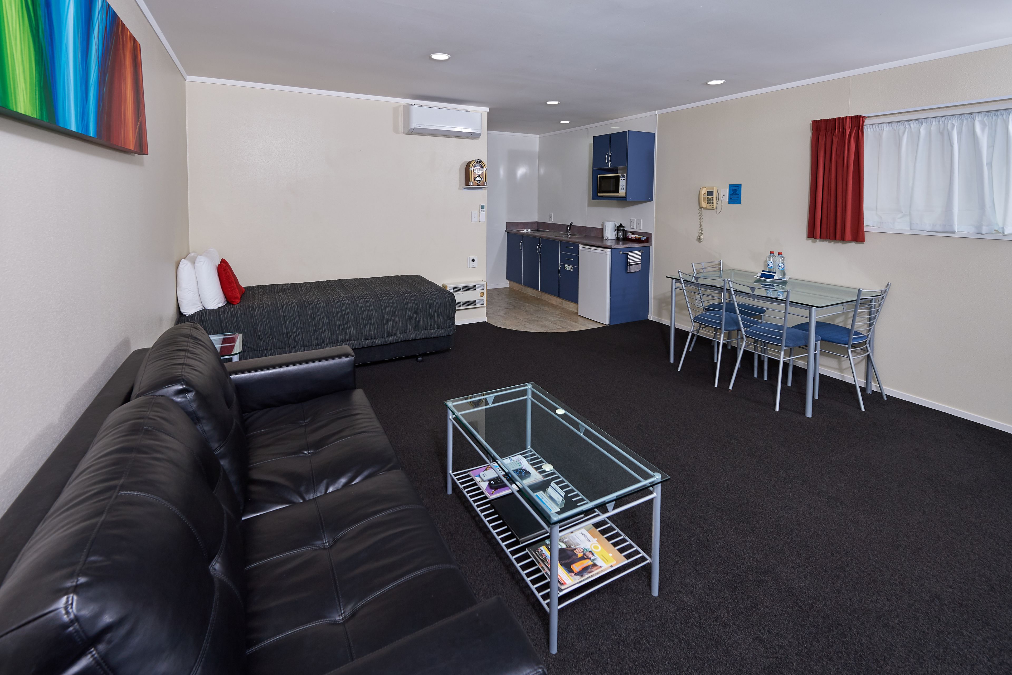 Una habitación compacta con cama, una mesita de cristal con revistas, kitchenette y zona de comedor con sillas.