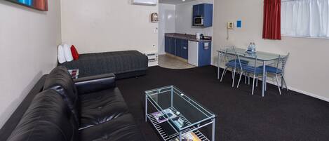 Una habitación compacta con cama, una mesita de cristal con revistas, kitchenette y zona de comedor con sillas.