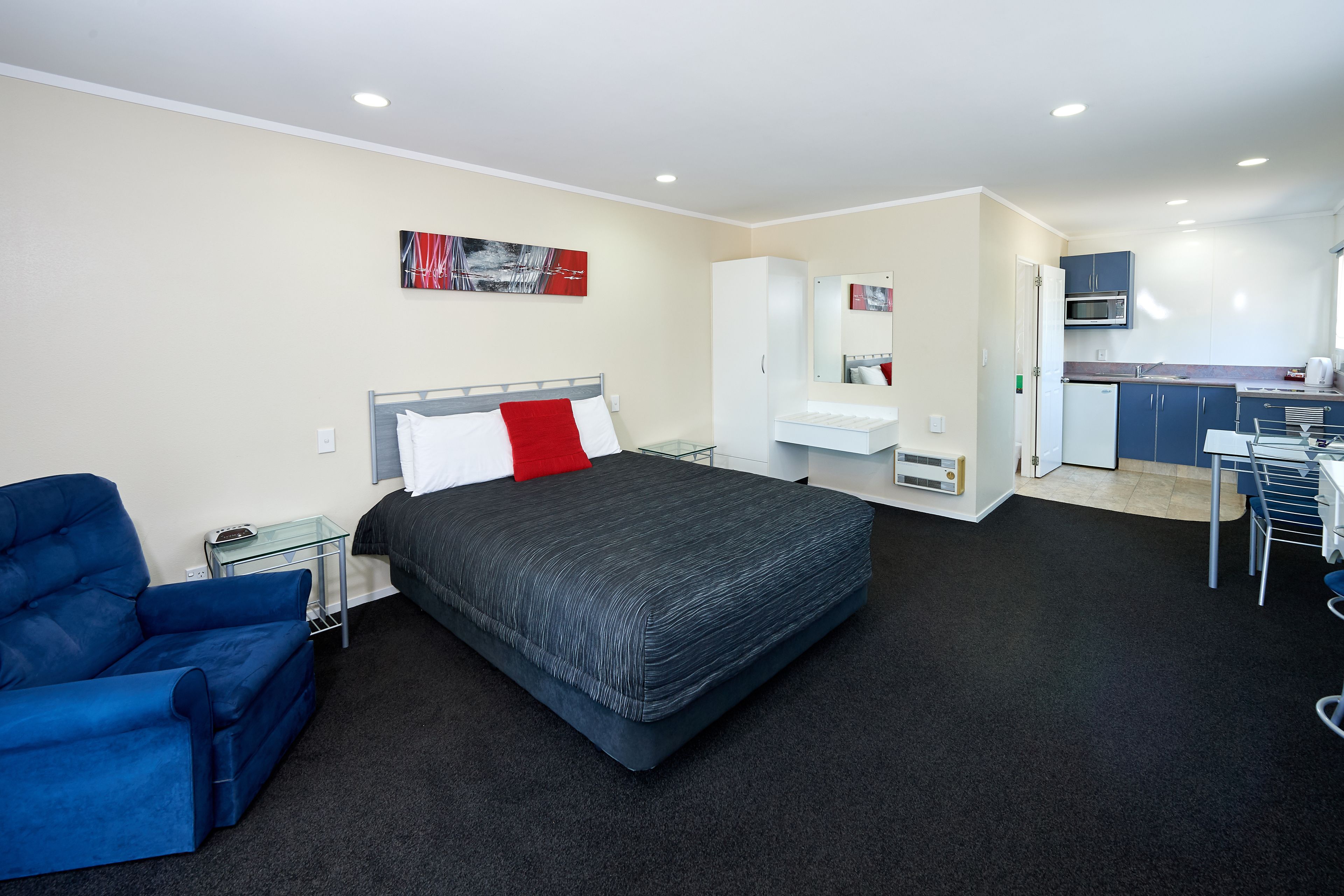 Una habitación de hotel moderna con una cama grande, un sillón azul, una kitchenette y un comedor.