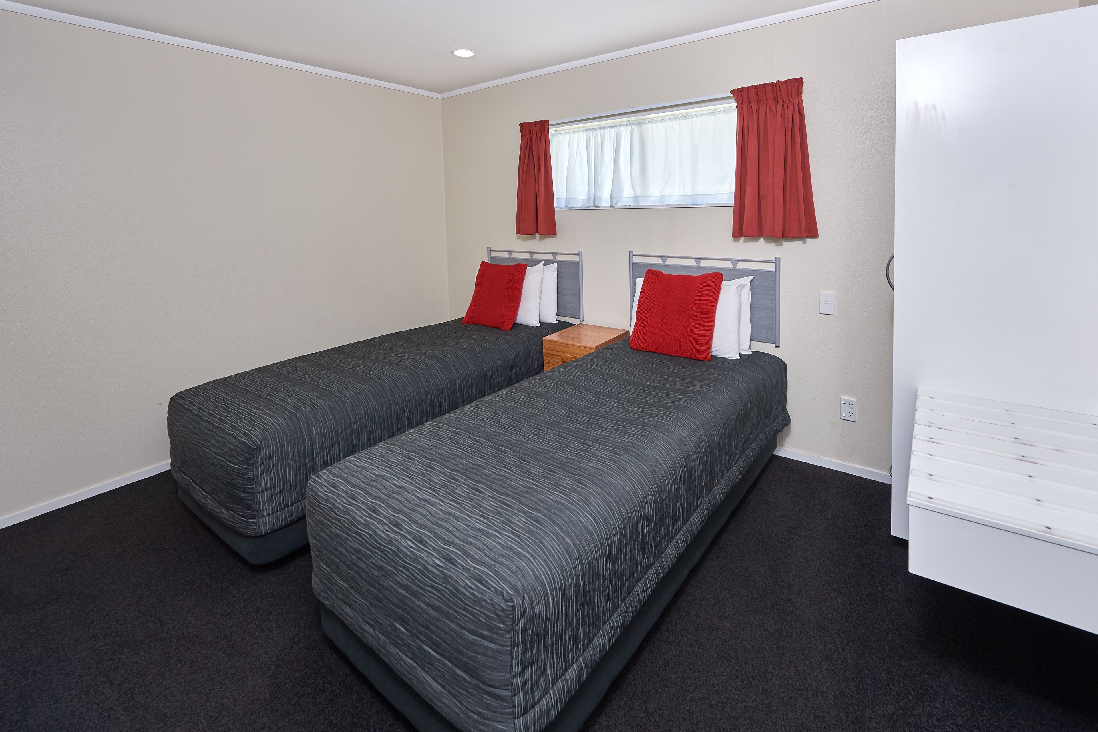 A room with two single beds, a wooden nightstand, a white wardrobe, and red curtains.