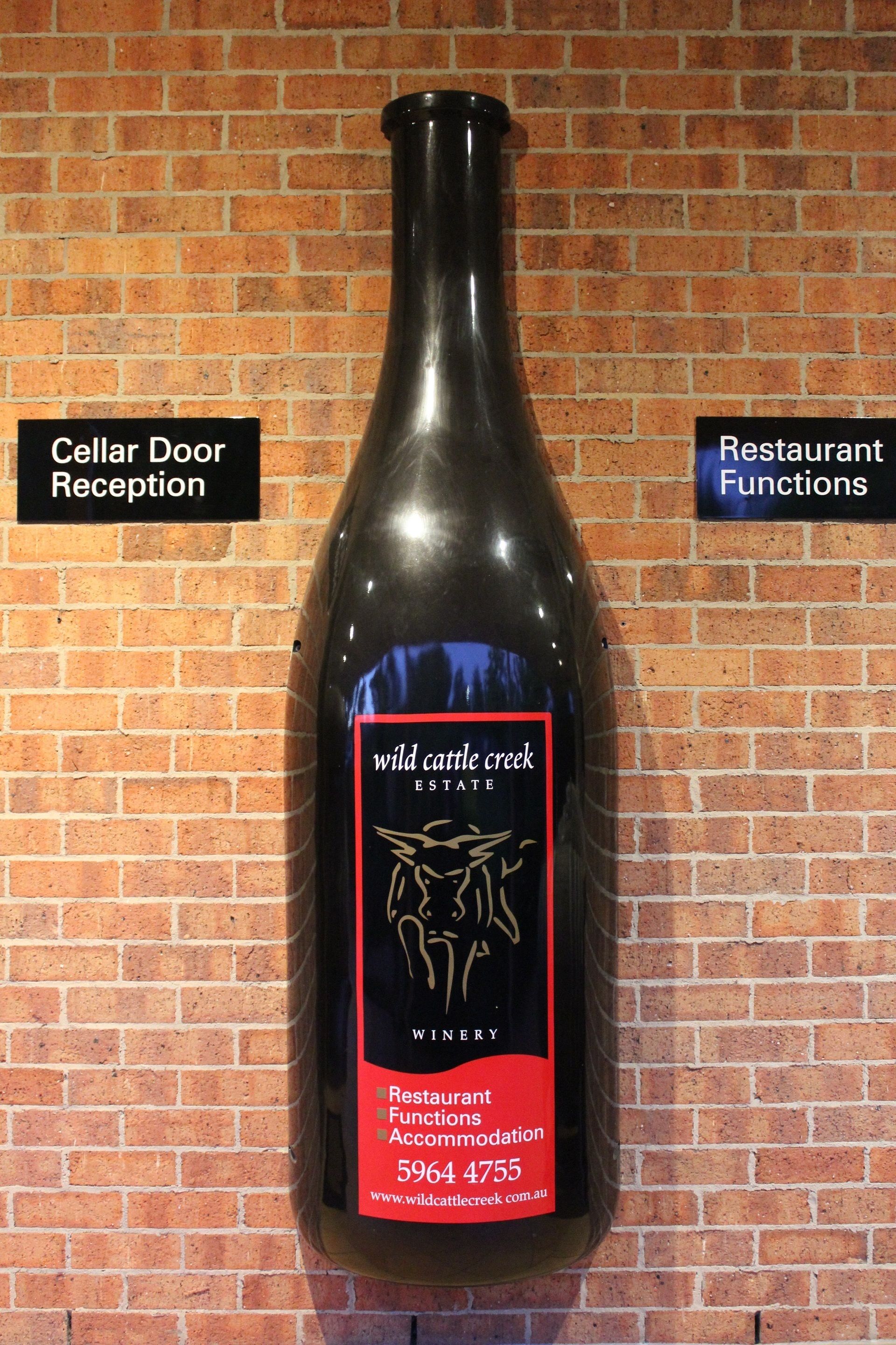 Une bouteille de vin portant l’étiquette « Wild Cattle Creek Estate » est posée contre un mur de briques, avec des panneaux indiquant « Accueil à la porte de la cave » et « Événements au restaurant ».