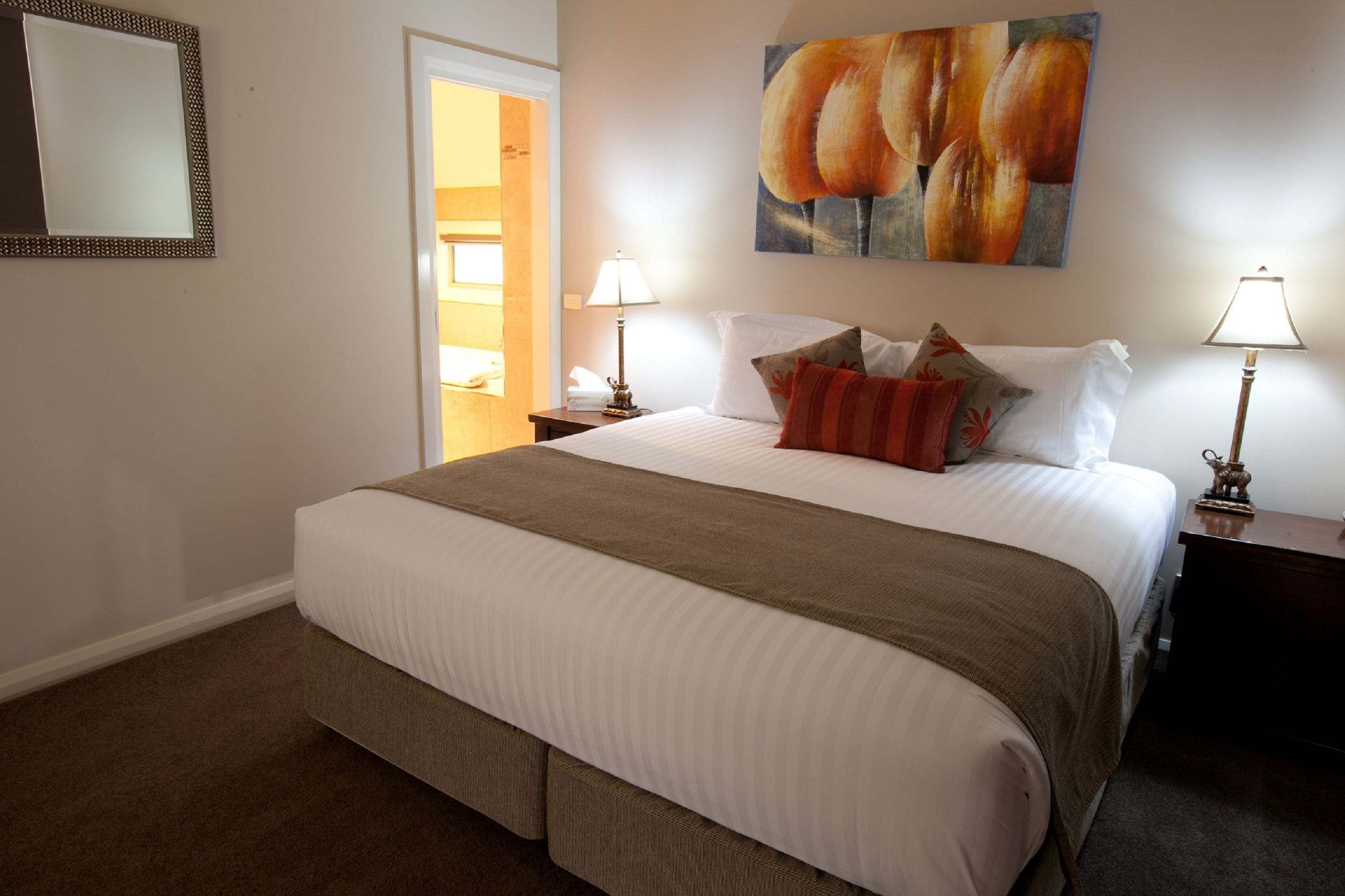 A neatly made bed with a brown blanket, two bedside lamps, and a painting of bread on the wall.