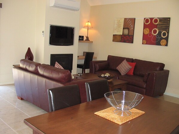 Un salón con un sofá de cuero marrón, una mesa de comedor con un centro de mesa de metal y un televisor montado en la pared.