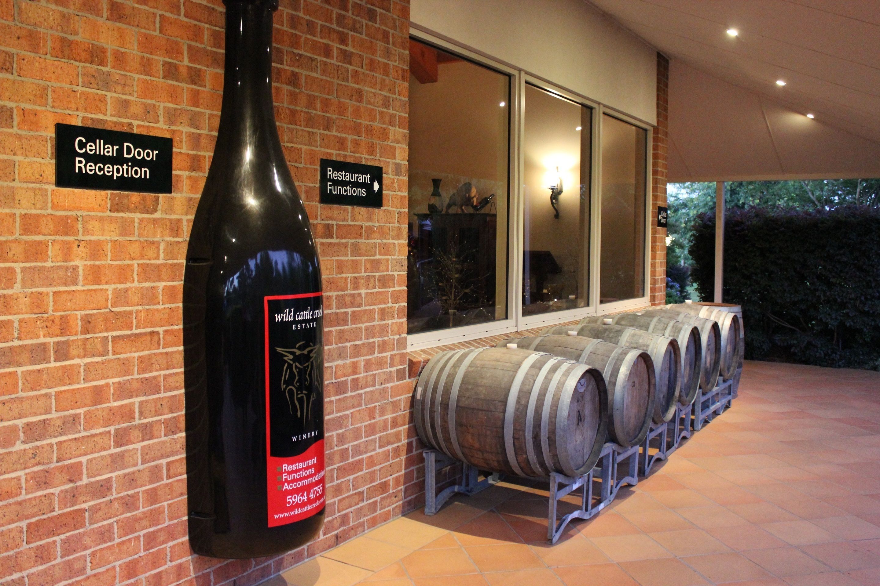 A brick building with a large wine bottle and several wooden barrels. Signs indicate a cellar door reception and restaurant functions.