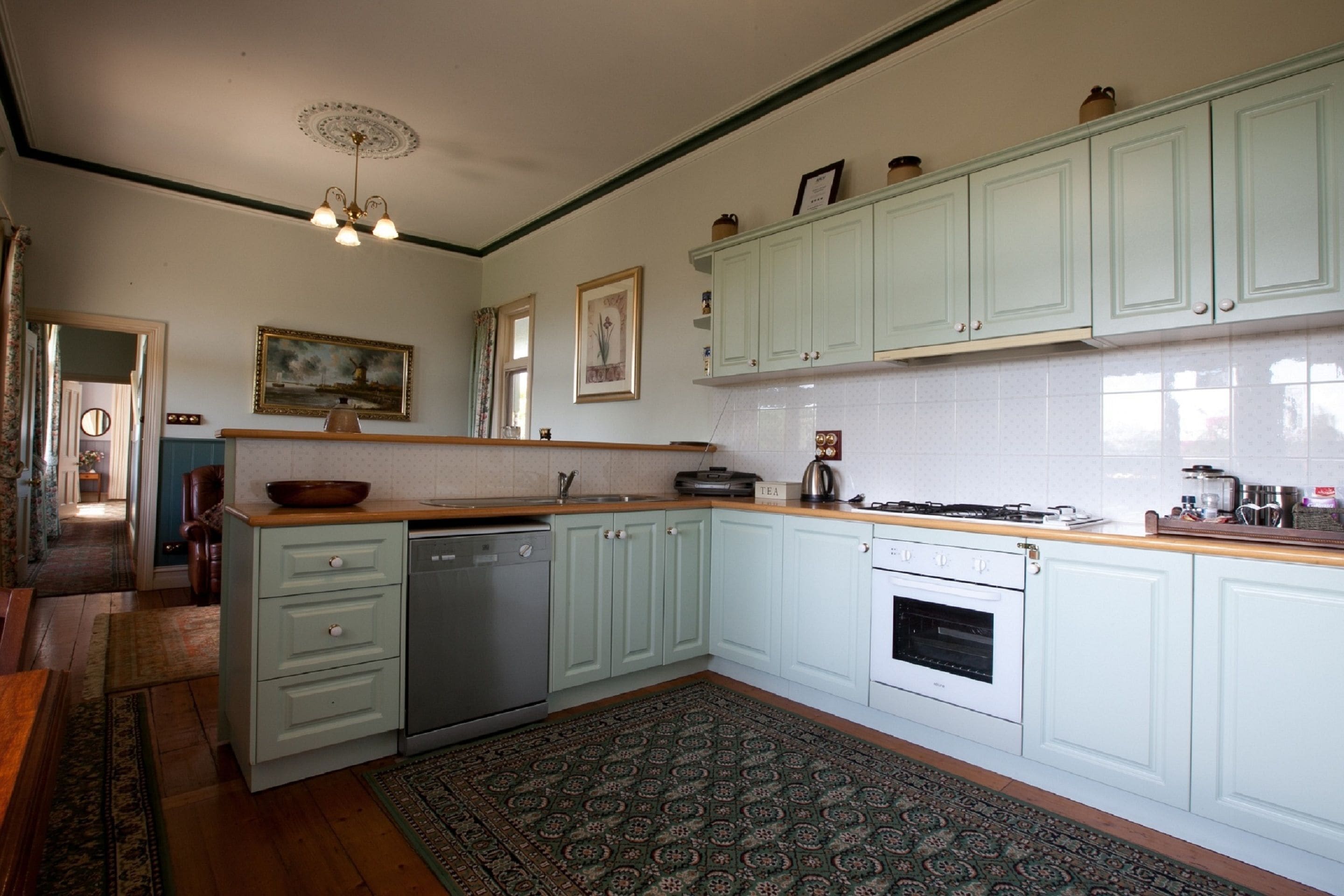 A kitchen with light green cabinets, a wooden countertop, a stove, and a dishwasher. There is a patterned rug on the floor.