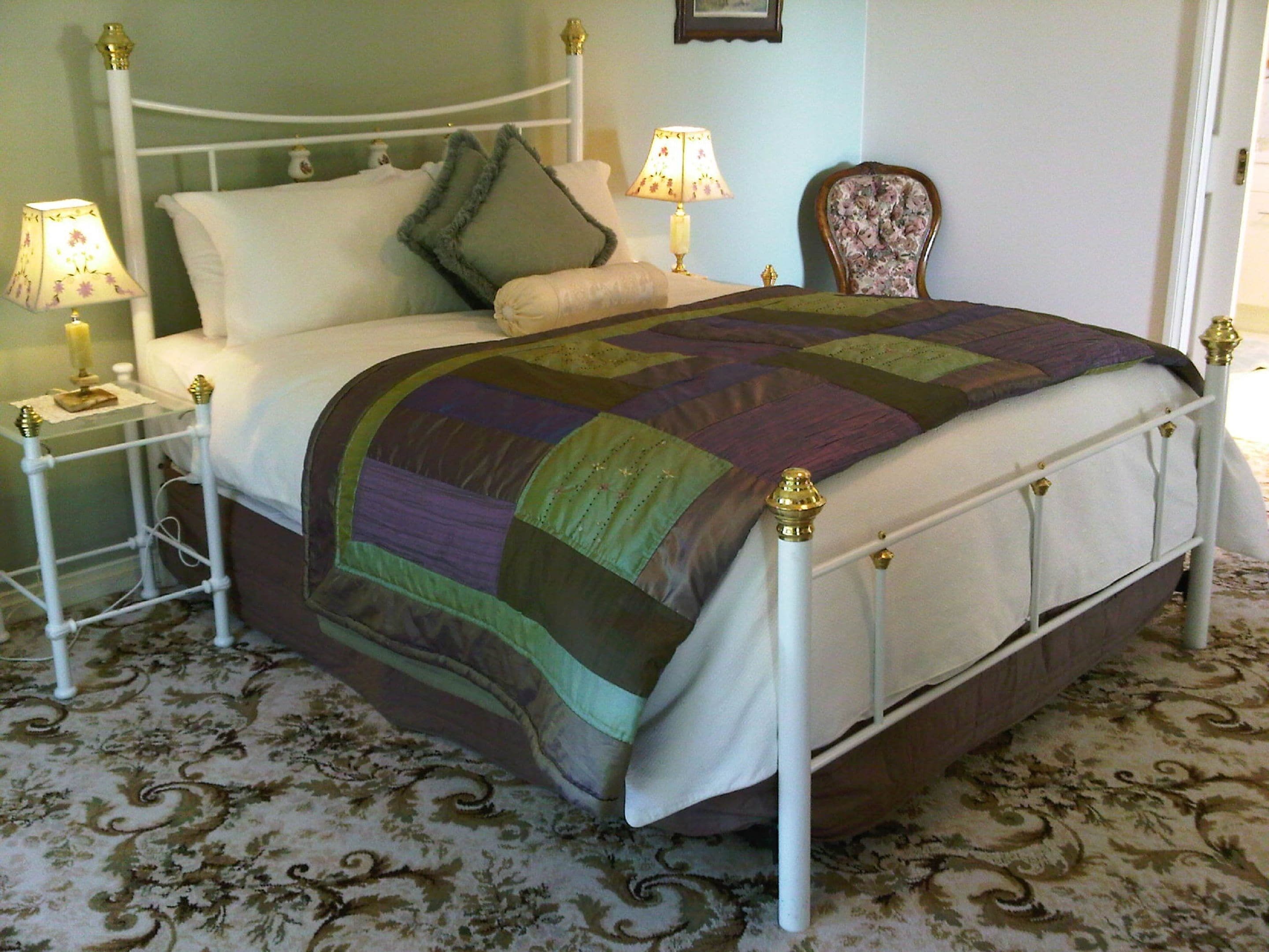 A neatly made bed with a colorful quilt, two bedside tables with lamps, a floral chair, and a patterned carpet.