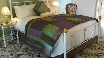 A neatly made bed with a colorful quilt, two bedside tables with lamps, a floral chair, and a patterned carpet.