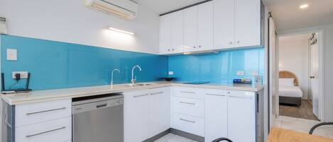 Eine kompakte Küche mit weißen Schränken, einer blauen Spritzschutzleiste, einem Geschirrspüler und einem Essbereich mit einem Holztisch.