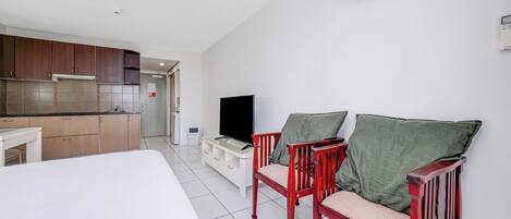 A hotel room with a kitchenette, a bed, two wooden chairs with cushions, a television, and an air conditioning unit.