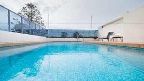 A clear blue pool with tiled edges, surrounded by a white wall and a glass fence. There are two lounge chairs on the right side.