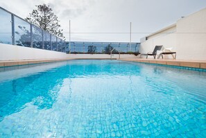 A clear blue pool with tiled edges, surrounded by a white wall and a glass fence. There are two lounge chairs on the right side.