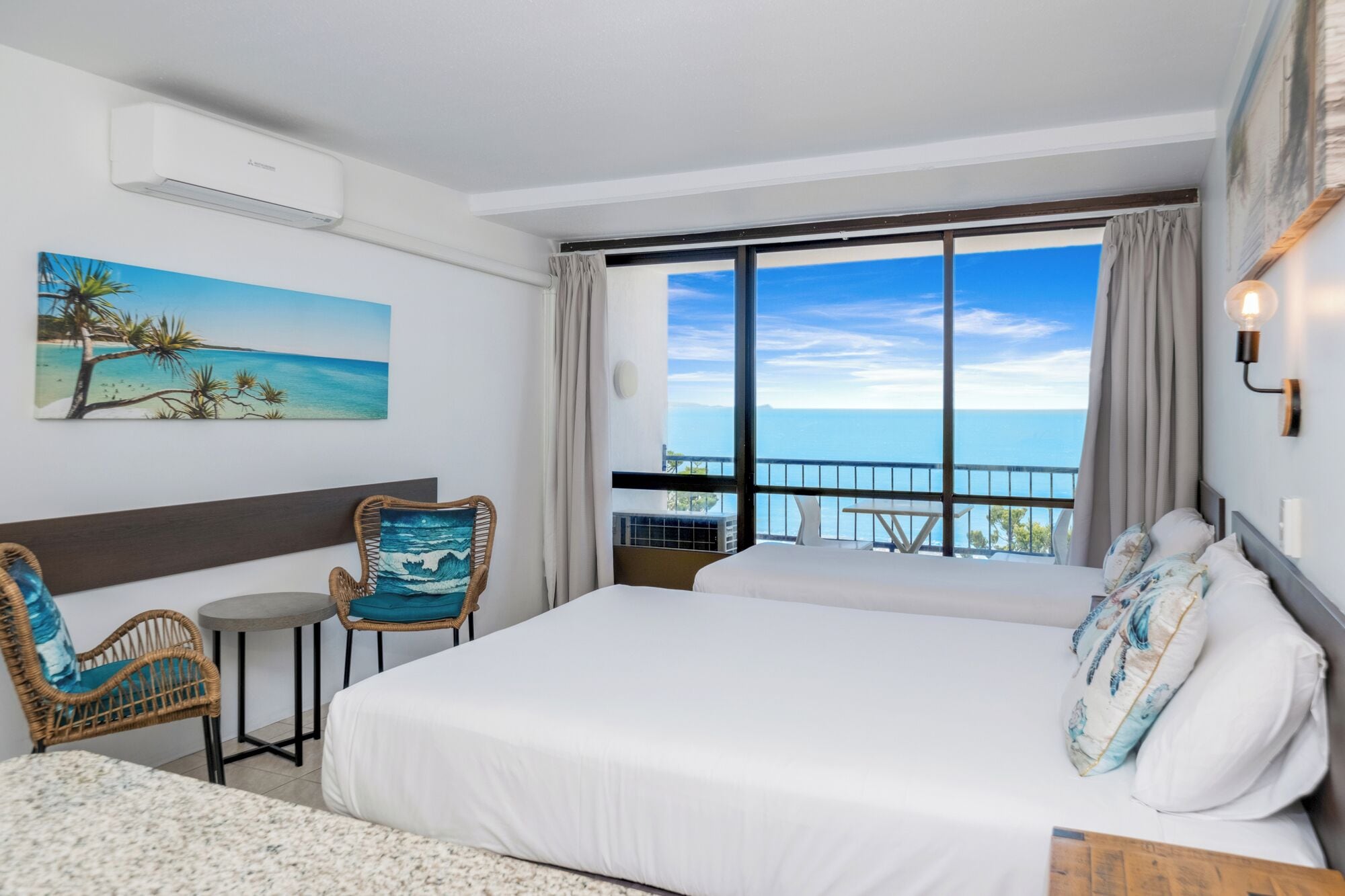 A hotel room with a bed, a chair, a small table, a painting of a beach, and a large window with a view of the sea.