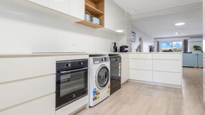 Une cuisine moderne avec des armoires blanches, un four encastré, un lave-linge et une étagère en bois au-dessus du four.