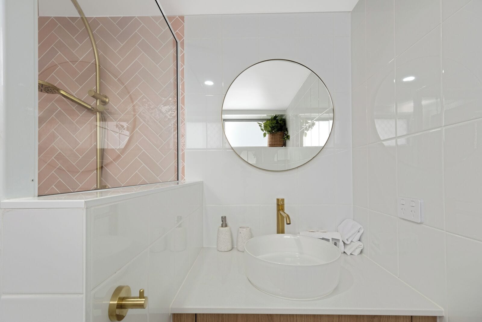 A modern bathroom with a glass-enclosed shower, a round mirror, and a white sink with gold fixtures.
