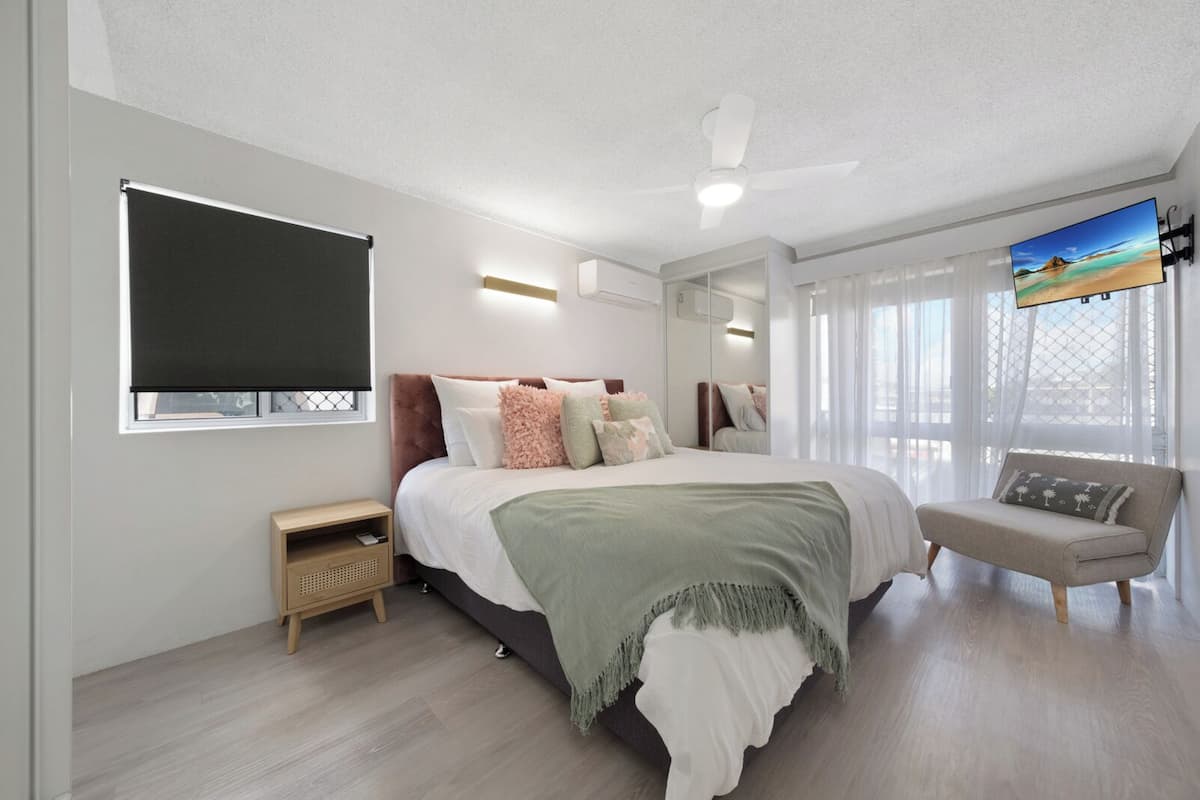Un dormitorio moderno con una cama grande, una mesita de noche, un sofá, un ventilador de techo y un televisor montado en la pared.