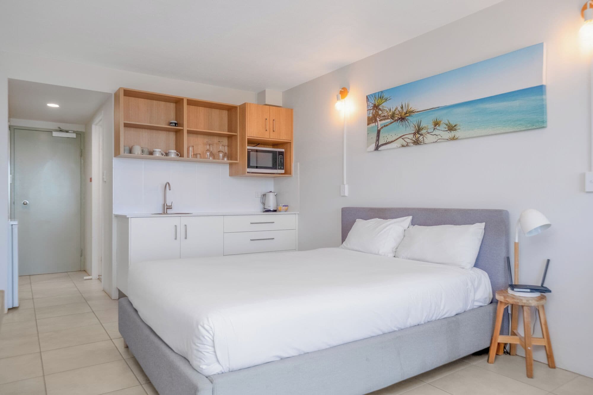 A bedroom with a bed, a small stool, a nightstand, a painting, and a kitchenette with wooden cabinets and a microwave.