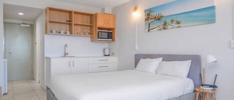 A bedroom with a bed, a small stool, a nightstand, a painting, and a kitchenette with wooden cabinets and a microwave.