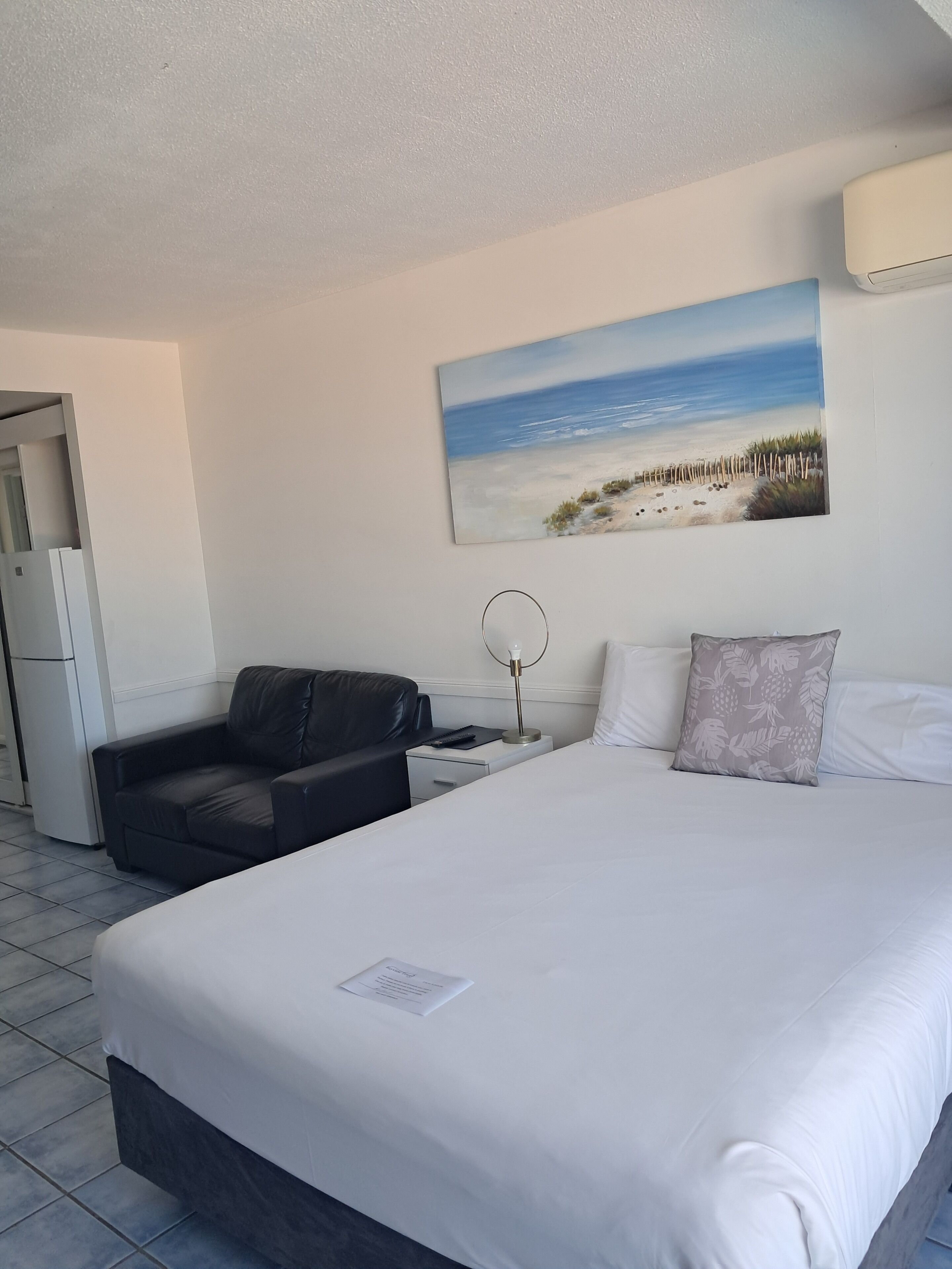 A hotel room with a bed, a black leather sofa, a desk with a lamp, a painting of a beach, and a white air conditioning unit.