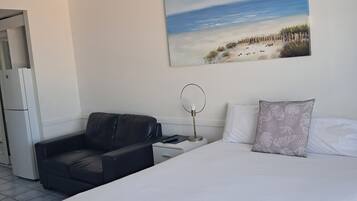 A hotel room with a bed, a black leather sofa, a desk with a lamp, a painting of a beach, and a white air conditioning unit.
