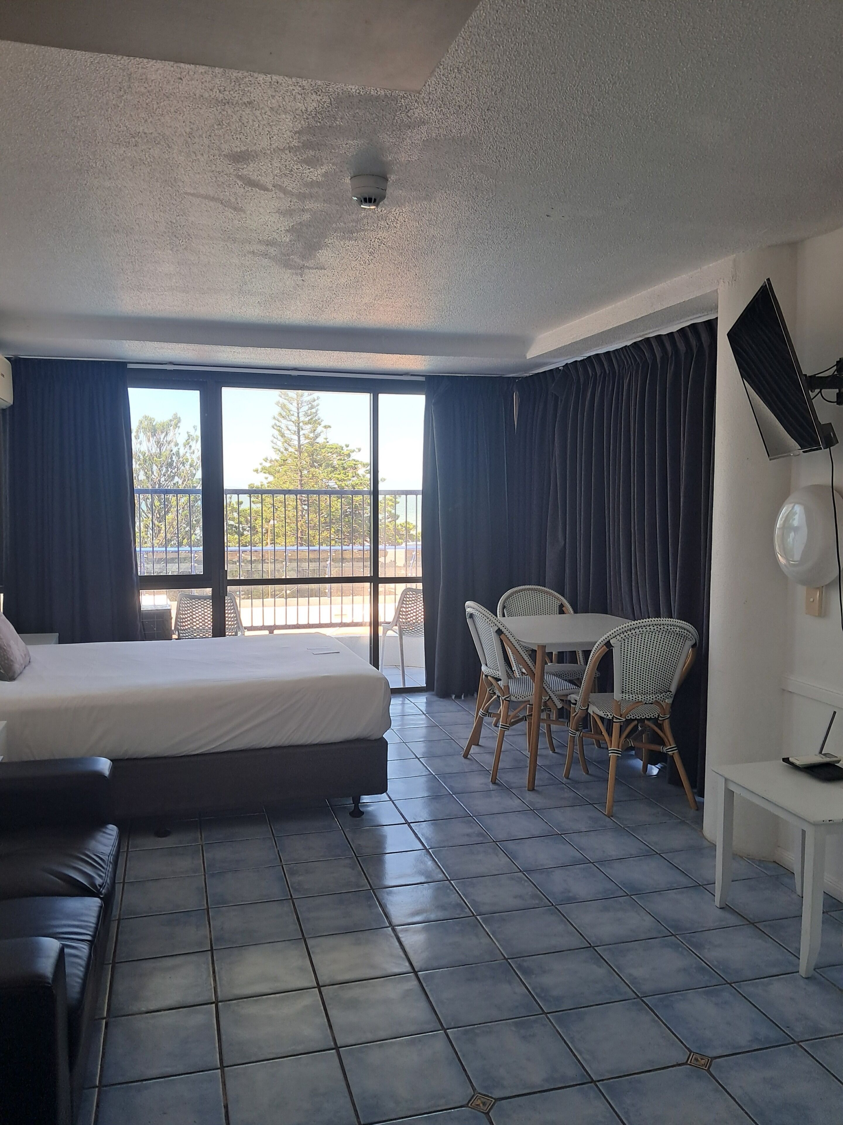 A hotel room with a bed, a dining table with chairs, a ceiling-mounted security camera, and a view of the outdoors.