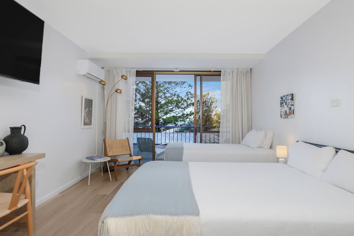Habitación de hotel con dos camas, un escritorio, una silla, un balcón con vistas y un cuadro en la pared.