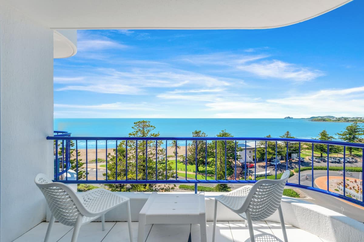 Un balcón con dos sillas blancas y una mesa, con vista a una playa y un cielo azul despejado.