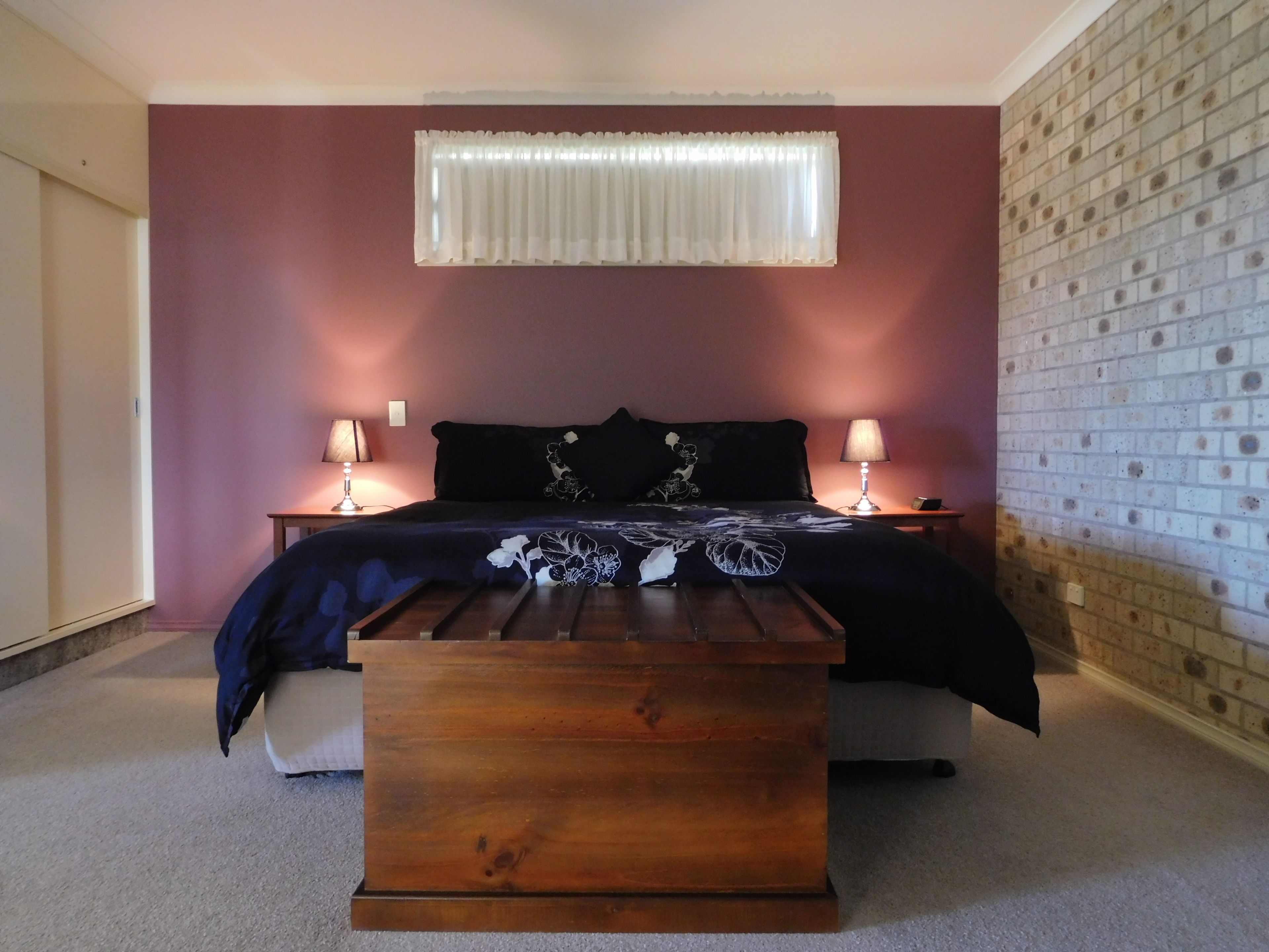 A bedroom with a bed, two bedside tables with lamps, a wooden bench, and a brick-patterned wall.