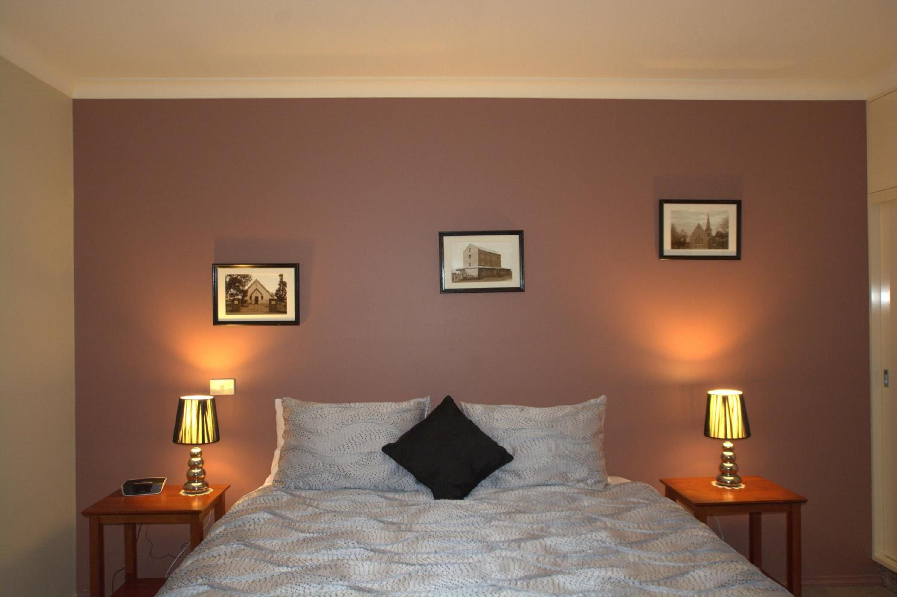 Una cama bien hecha con una colcha estampada y dos almohadas, flanqueada por mesitas de noche con lámparas. Encima de la cama, hay tres cuadros enmarcados en la pared.