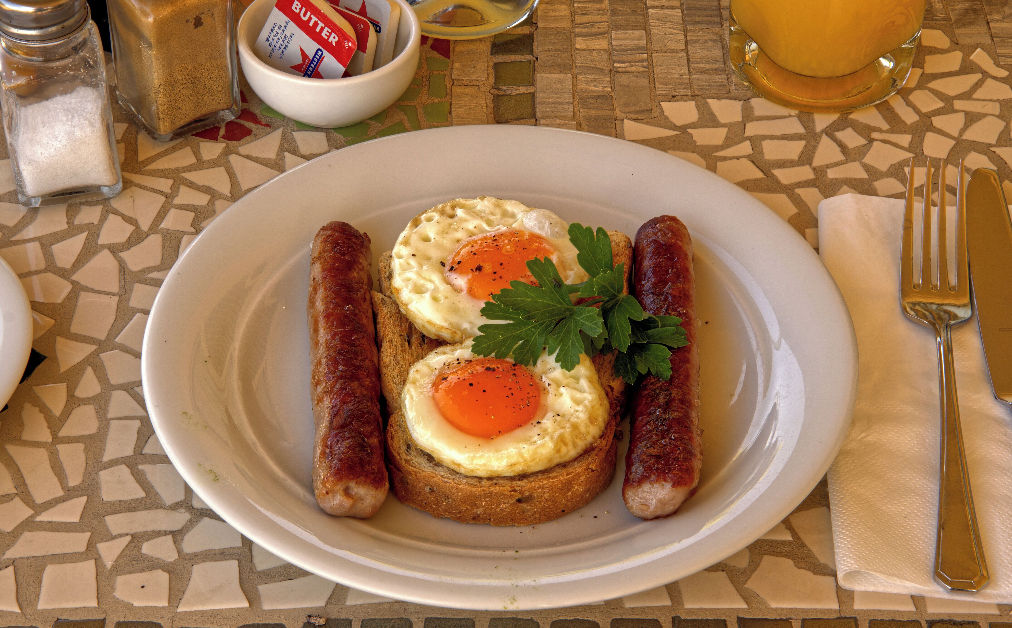 Une assiette contenant deux saucisses, deux œufs au plat et une garniture, le tout posé sur une table carrelée.