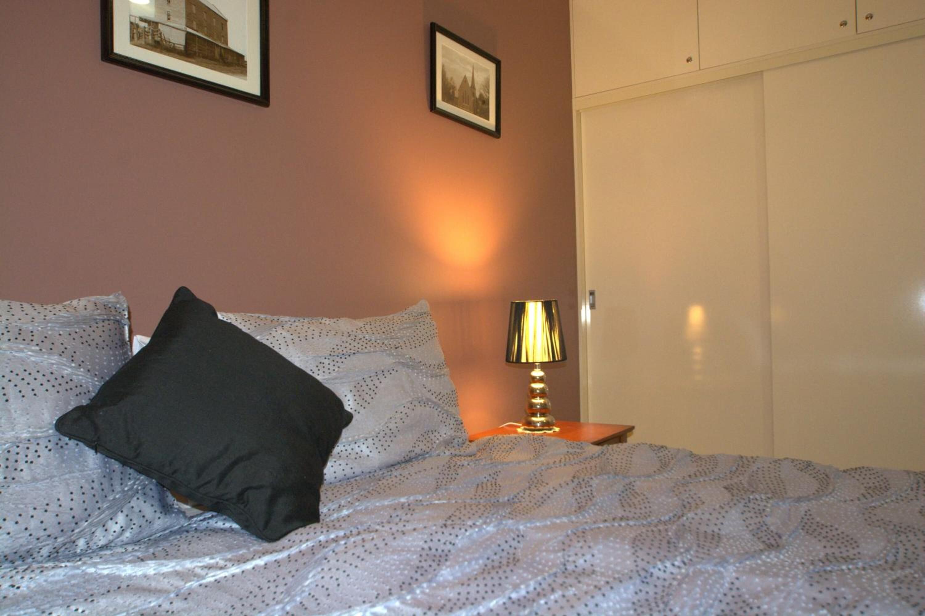 A neatly made bed with a patterned bedspread and two pillows, a bedside lamp, and a wardrobe in a room with a warm wall color.