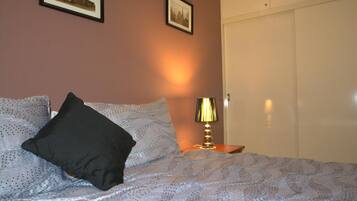 A neatly made bed with a patterned bedspread and two pillows, a bedside lamp, and a wardrobe in a room with a warm wall color.