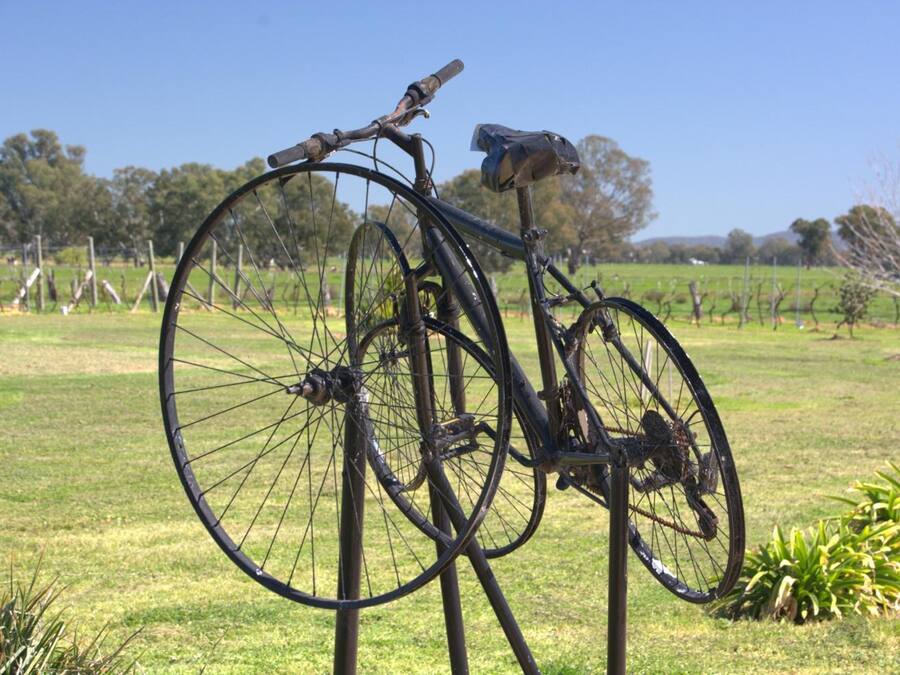 A bicycle leaning against a metal post in a grassy field with trees and a fence in the background.