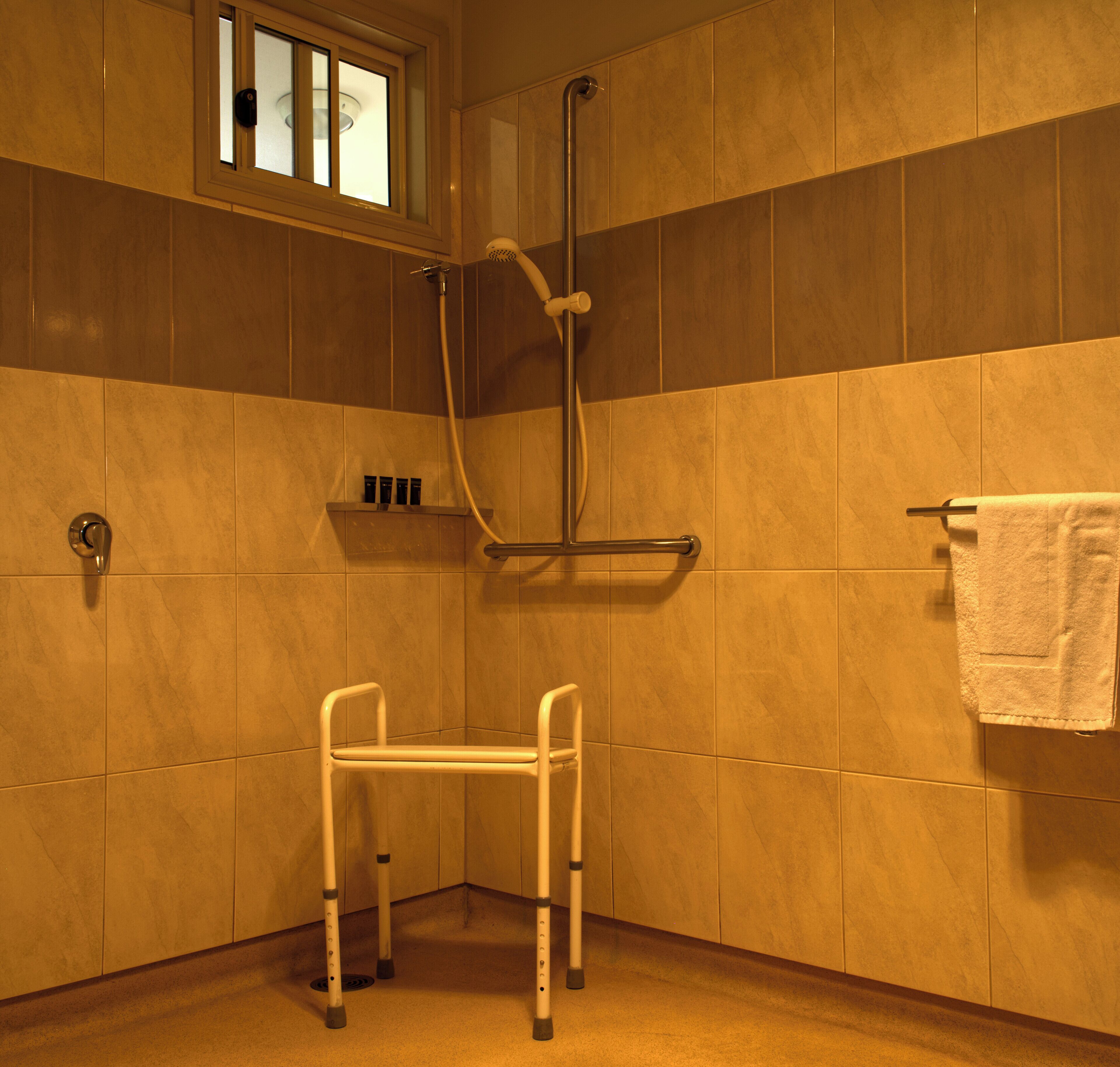 Zona doccia con sedia per disabili, porta asciugamani e finestra sopra la doccia.