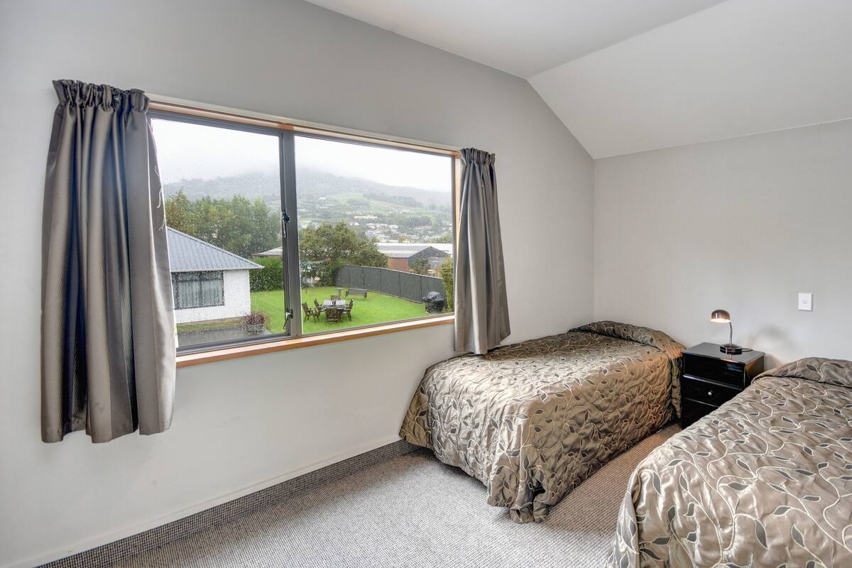 A bedroom with two beds, a window with curtains, a bedside table with a lamp, and a view of a green yard and buildings.