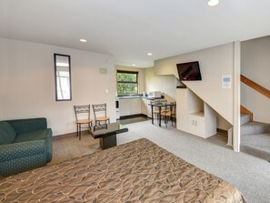 A compact room with a sofa, chairs, a small table, a bed, a kitchenette, and a TV.