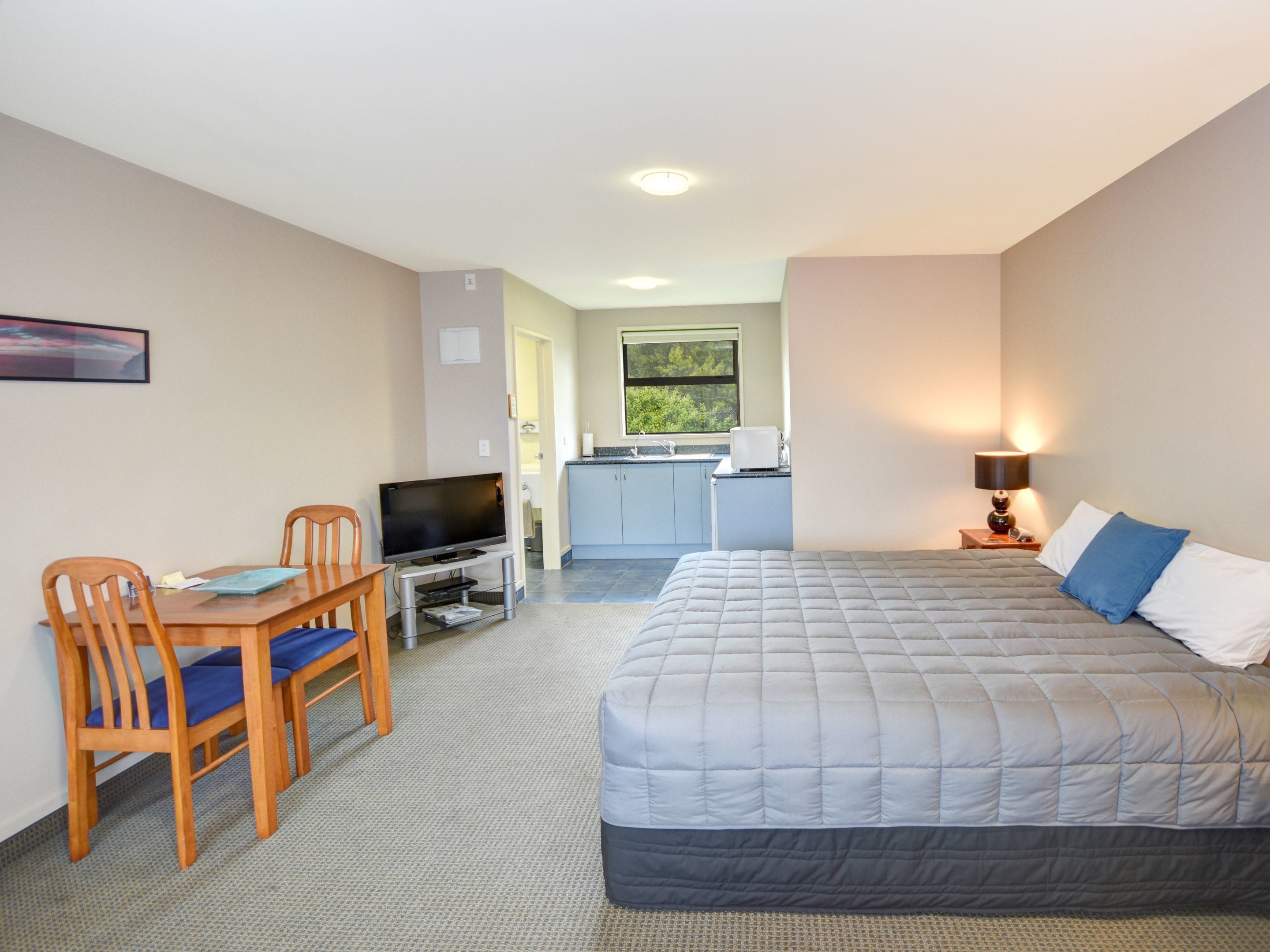 A bedroom with a bed, a desk with a TV, a chair, and a kitchenette in the background.