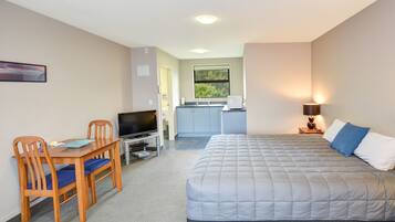 A bedroom with a bed, a desk with a TV, a chair, and a kitchenette in the background.