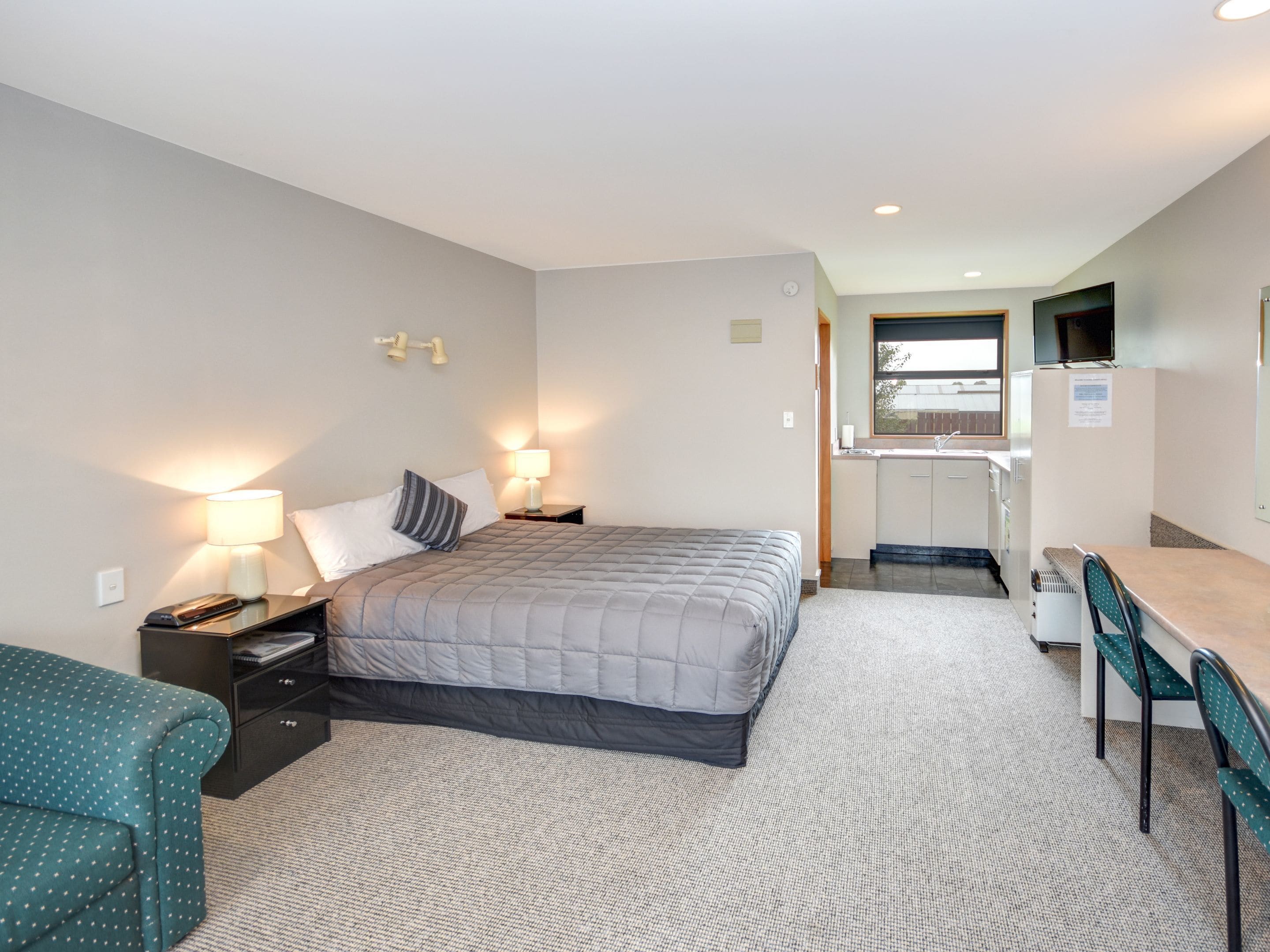 A compact hotel room with a bed, bedside tables, a desk, and a kitchenette.
