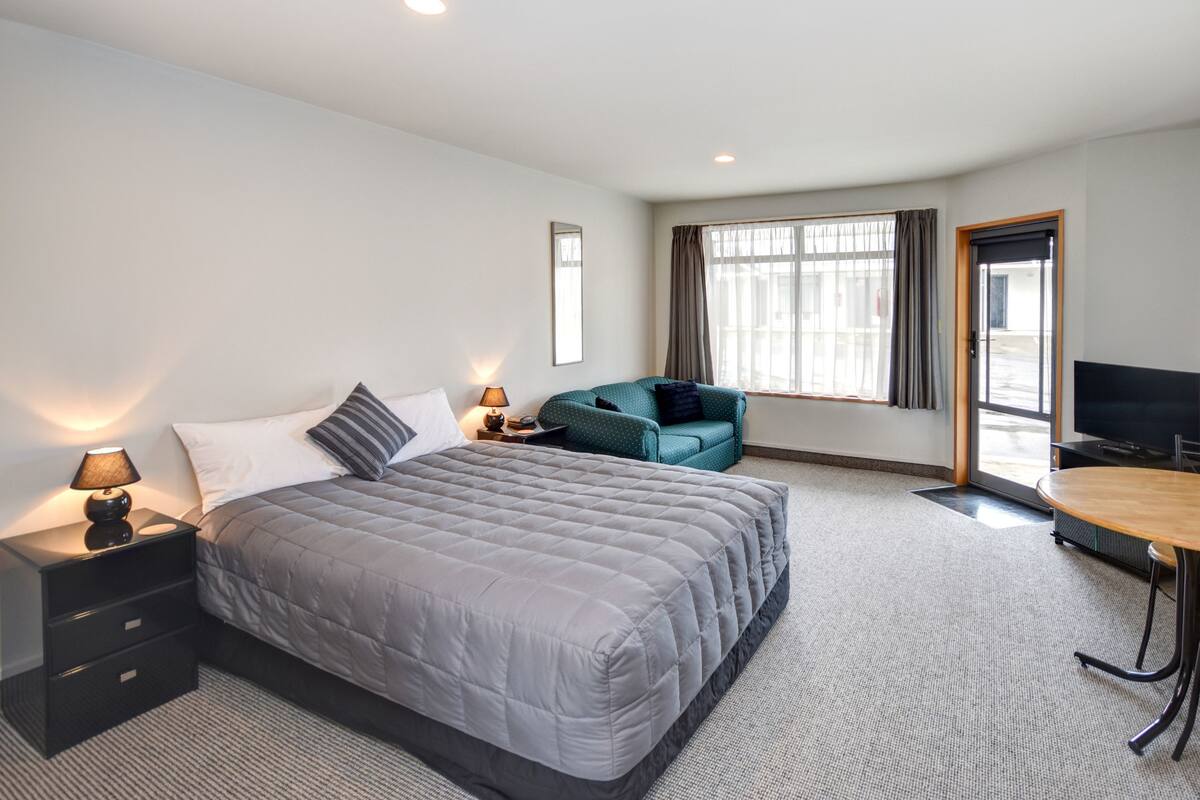 A hotel room with a large bed, a desk, a chair, a TV, and a window with curtains.