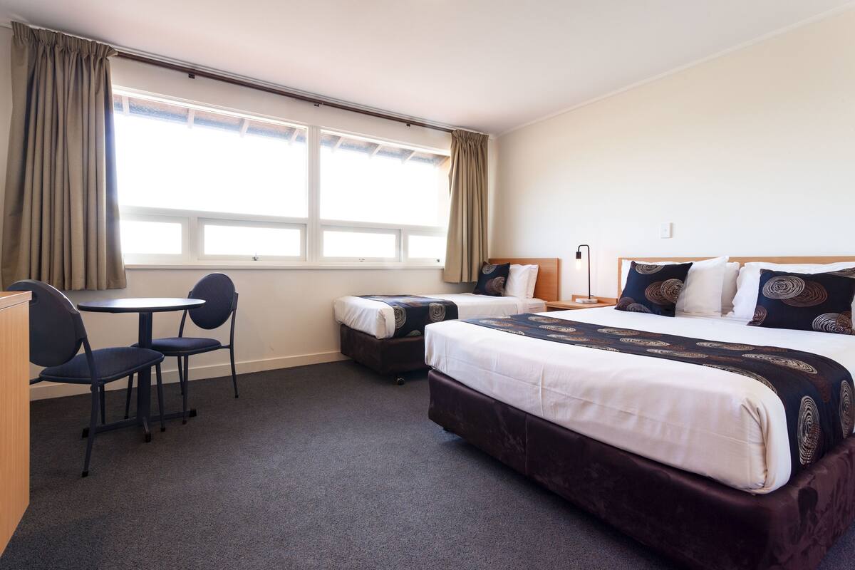 Quarto de hotel com cama, duas mesas de cabeceira, escrivaninha com cadeira e janela com cortinas.