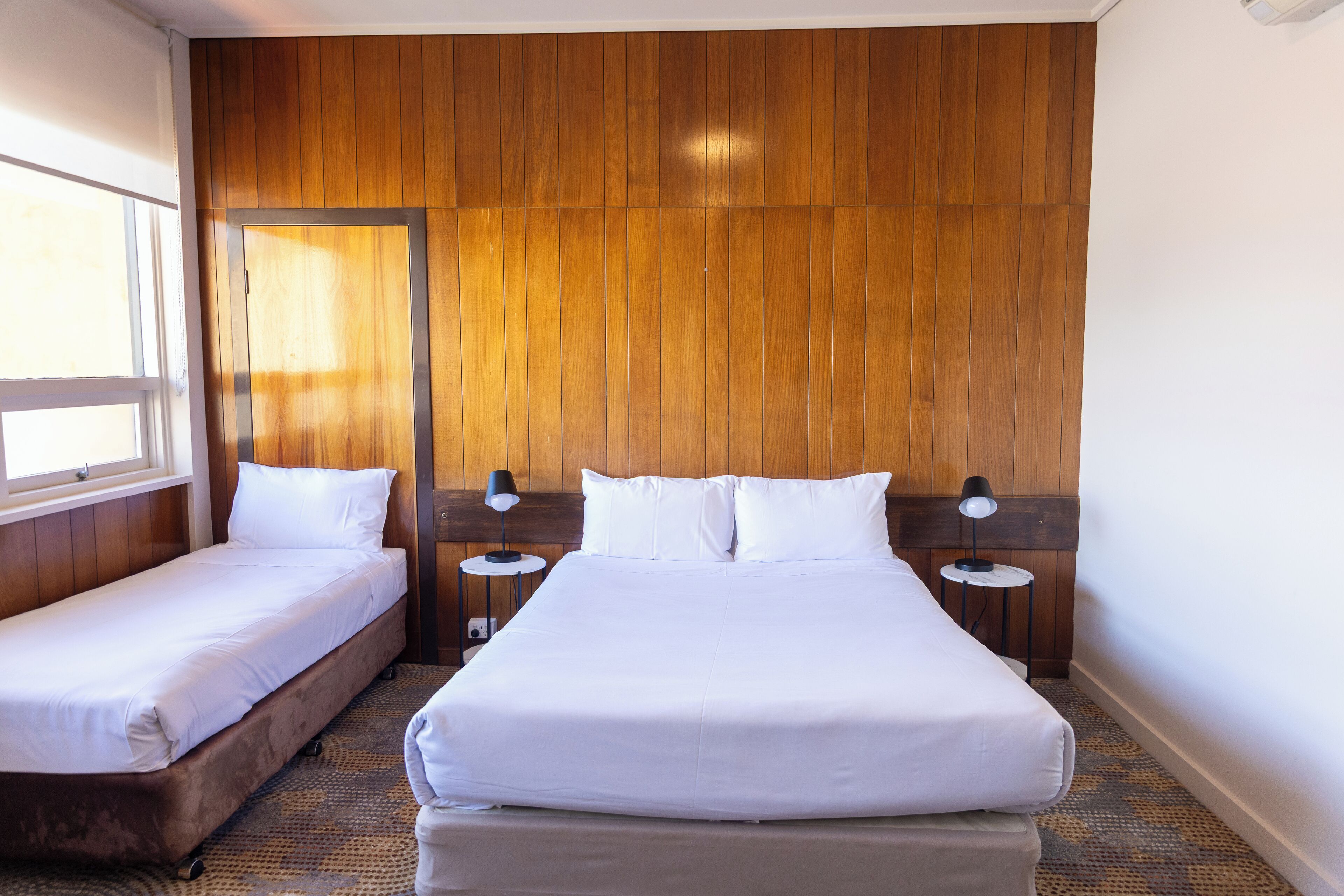 Habitación de hotel con dos camas individuales, un panel de pared de madera, una ventana y dos mesitas de noche con lámparas.