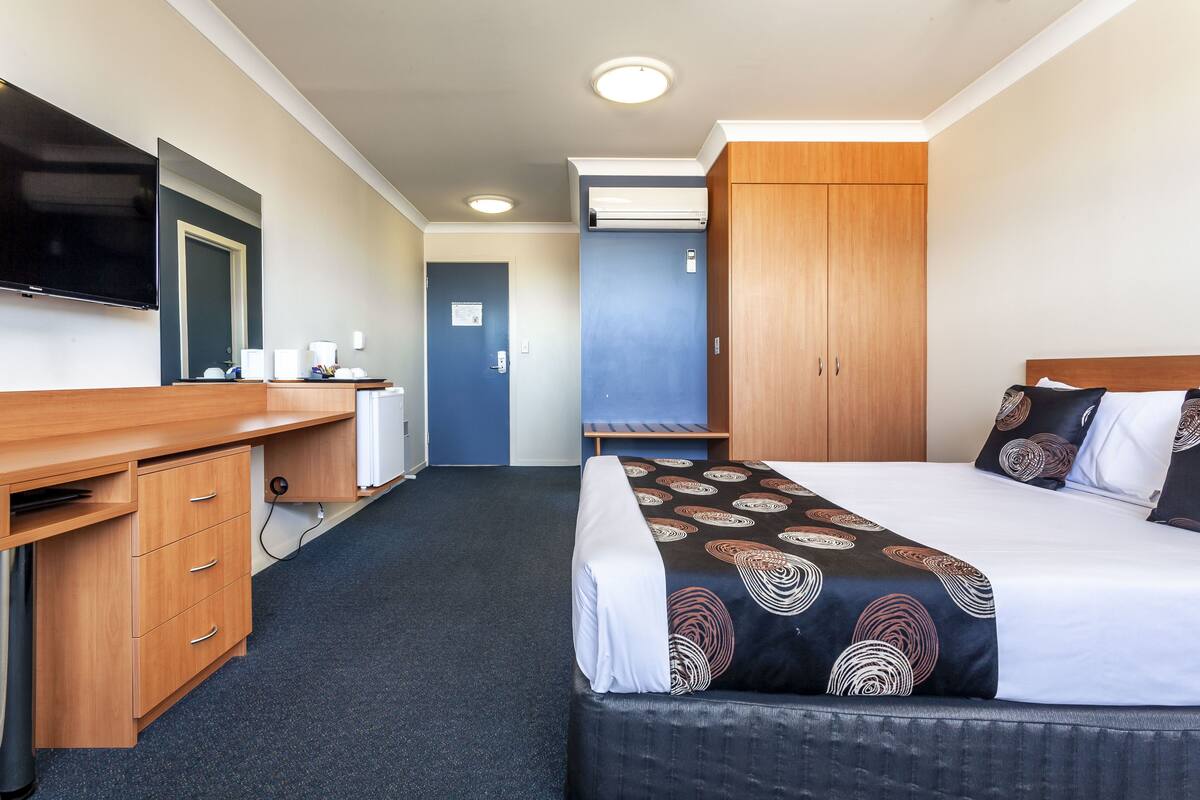 Quarto de hotel com cama, escrivaninha com gavetas, televisão e guarda-roupa.