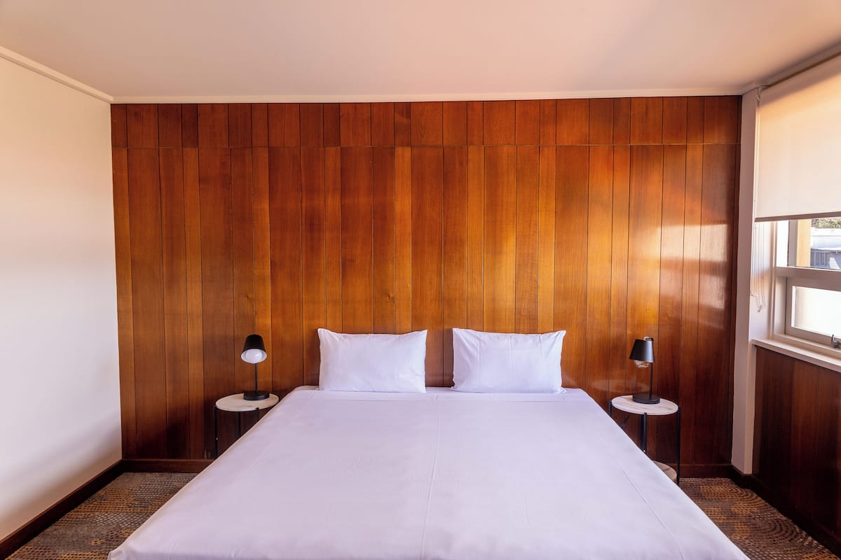 Quarto de hotel com cabeceira de madeira, uma cama grande com roupa de cama branca, duas mesas de cabeceira com luminárias e uma janela com persianas.
