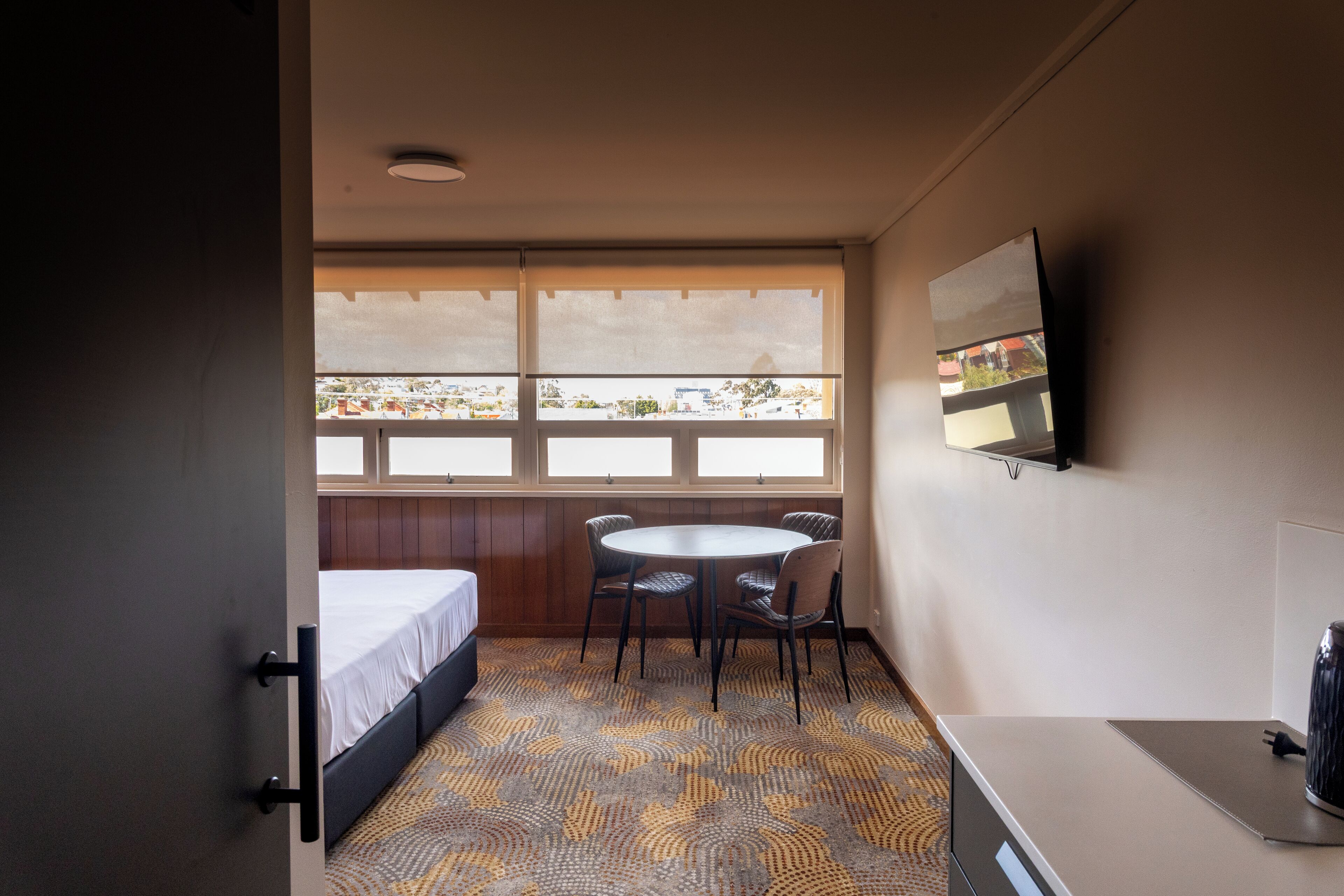 A hotel room with a bed, a small table with chairs, a TV on the wall, and a window with a view of buildings.