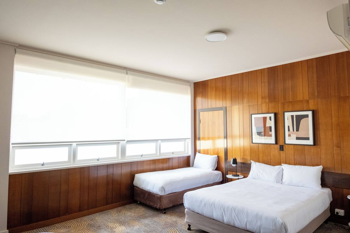 Quarto de hotel com uma janela grande, duas camas, painéis de madeira e dois quadros na parede.