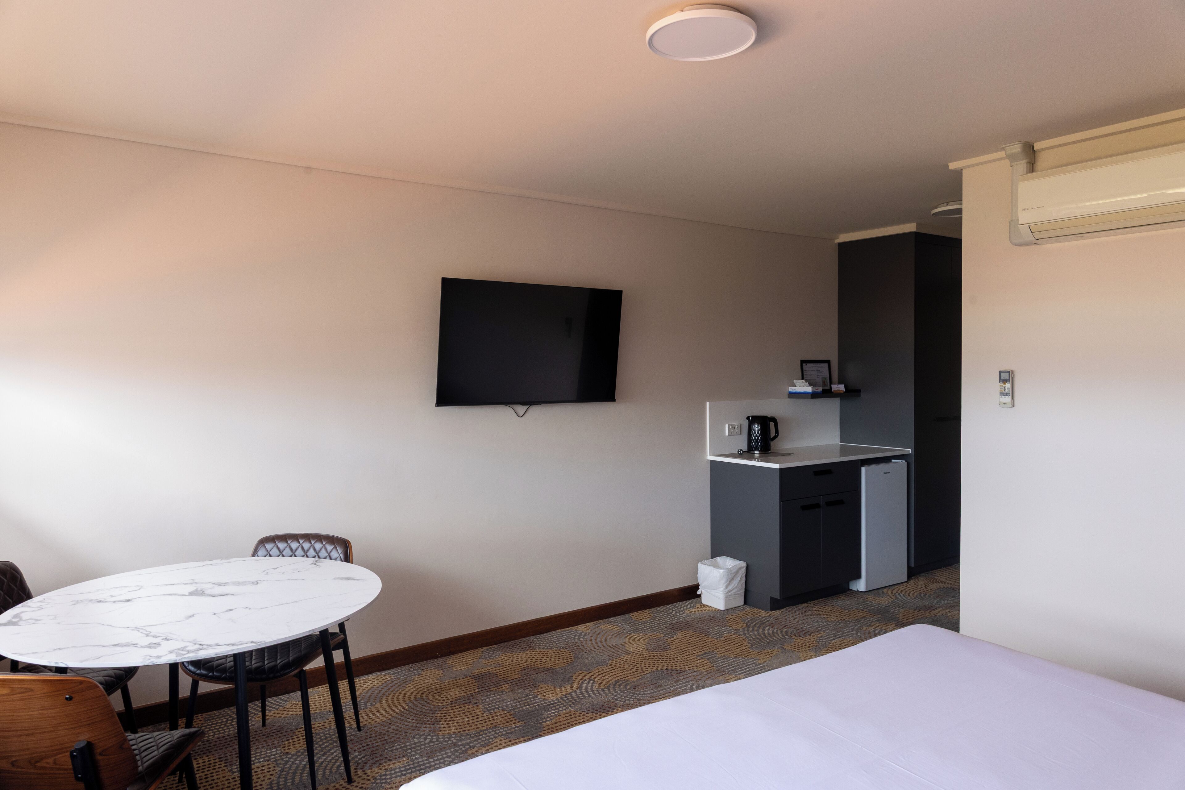 Una camera d'albergo con un letto, un tavolino rotondo con sedie, una televisione a parete, un angolo cottura con una macchina da caffè e un cestino.