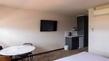 Una camera d'albergo con un letto, un tavolino rotondo con sedie, una televisione a parete, un angolo cottura con una macchina da caffè e un cestino.