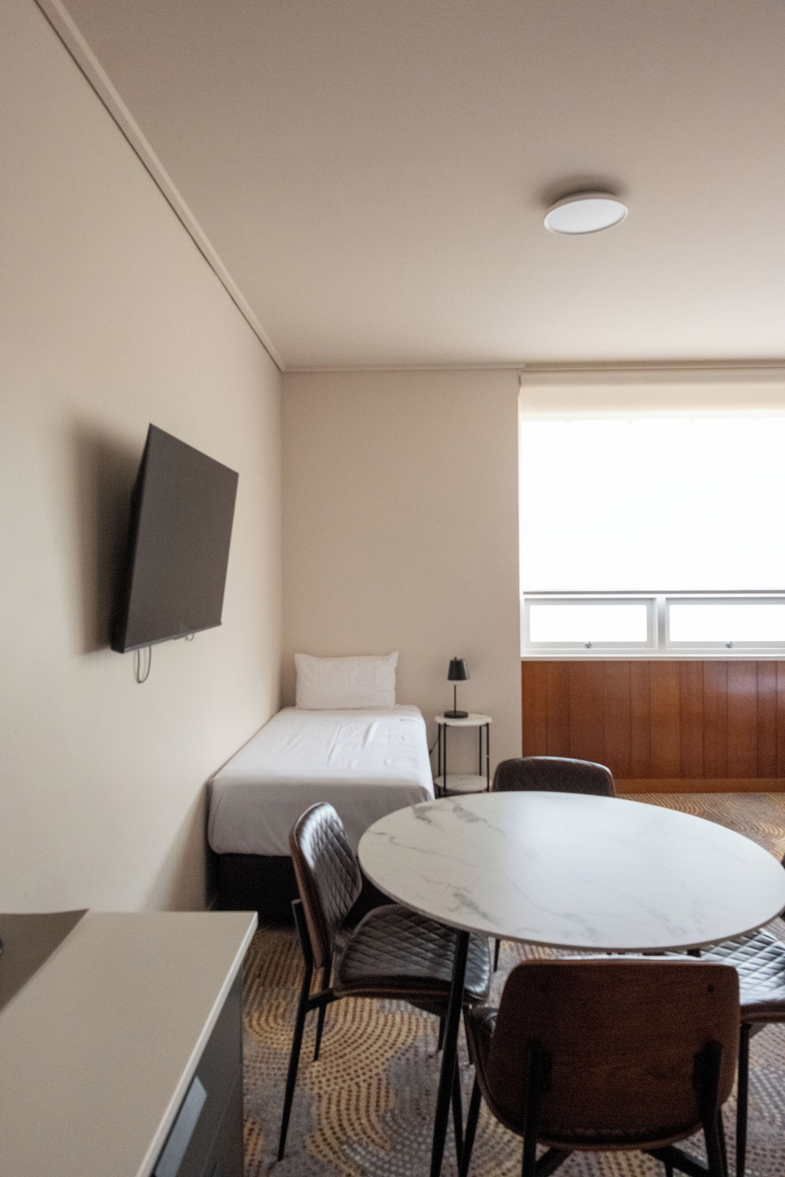 A hotel room with a bed, a TV mounted on the wall, a round table with chairs, and a window providing natural light.