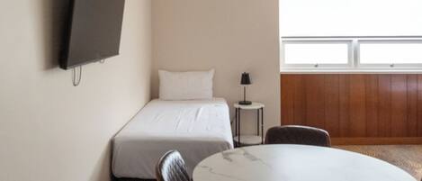 A hotel room with a bed, a TV mounted on the wall, a round table with chairs, and a window providing natural light.