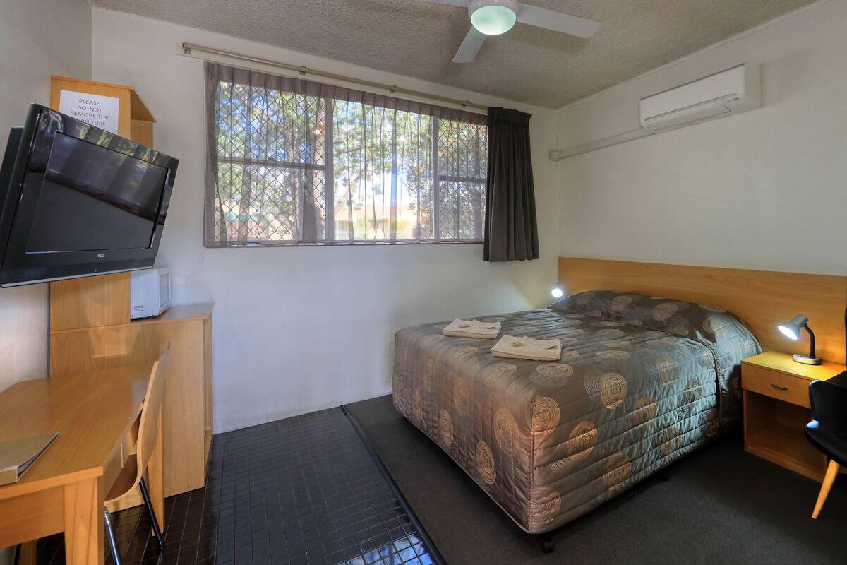 A bedroom with a bed, a desk, a chair, a TV, and a window with curtains.